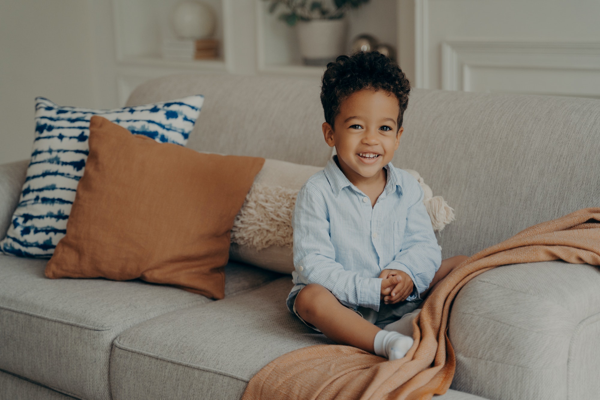 Sweet afro kid boy in casual clothes sitting on comfortable sofa and playing indoors